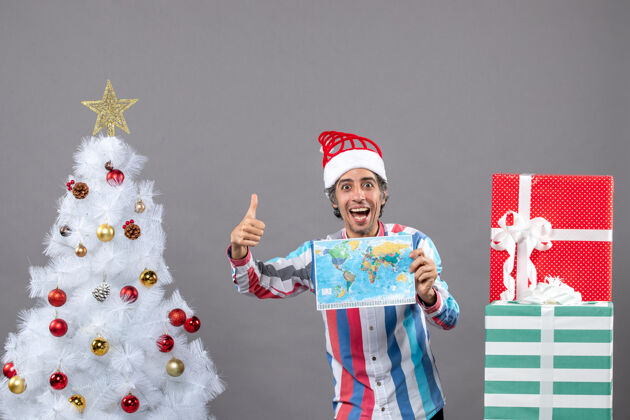 灰色正面图：快乐的男人戴着螺旋弹簧圣诞帽 竖起大拇指 举着世界地图家伙人向上
