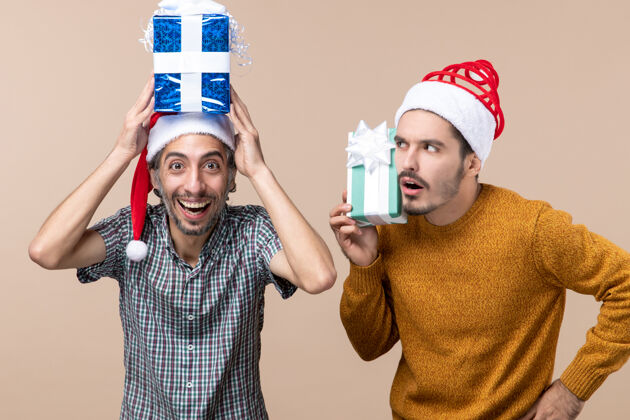 前面正面图两个戴着圣诞帽的家伙一个把礼物戴在头上 另一个在米色孤立背景上思考帽子两个视图