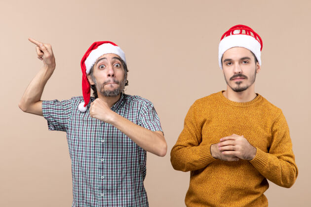 圣诞老人正面图两个戴着圣诞帽的家伙 其中一个站在米色孤立的背景上 表现出极大的兴趣男伙计们两个伙计们