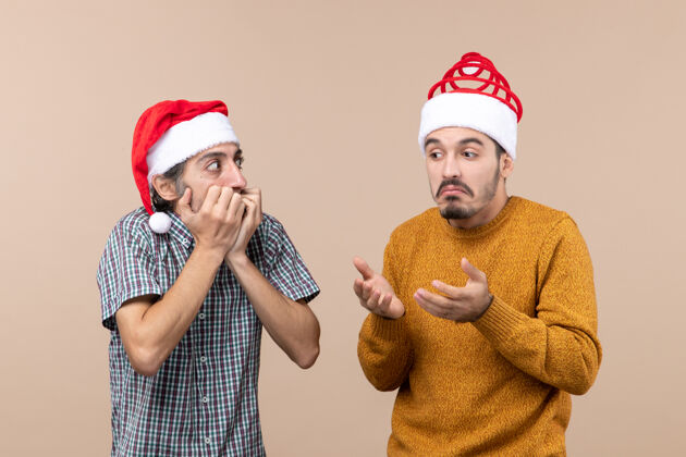 伙计们正面图：两个戴着圣诞老人帽子的男人在米色的孤立背景下大吃一惊两个惊奇的家伙男性帽子