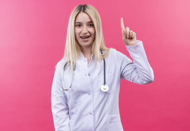 点微笑的医生年轻的金发女孩穿着医用长袍戴着听诊器和牙套指着孤立的粉红色背景听诊器粉红色牙科