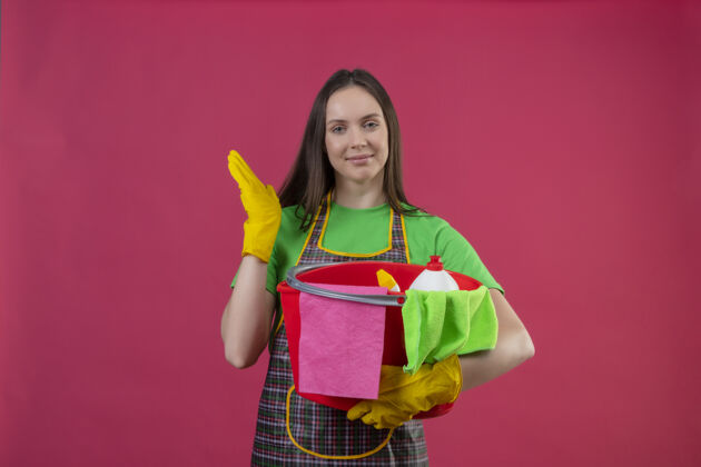 戴着高兴的清洁年轻女孩穿着制服 戴着手套 手持清洁工具 在孤立的粉红色背景上举手手套清洁手