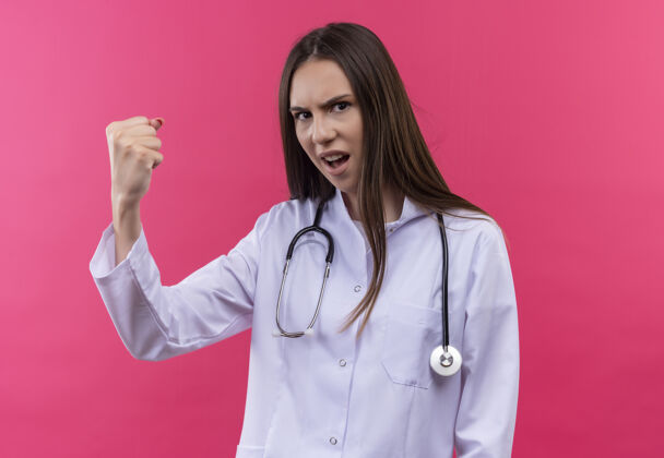 长袍严格的年轻医生女孩穿着听诊器医用长袍在孤立的粉红色背景上做强烈的手势女孩严格穿着