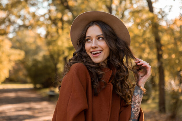 欢快画中有魅力的时尚微笑女子留着长长的卷发漫步在公园里穿着温暖的棕色外套秋日时尚 街头风格戴着帽子苗条休闲漂亮