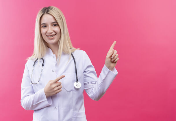 金发微笑的医生年轻的金发女孩穿着医用长袍戴着听诊器和牙套指向一边 双手放在孤立的粉红色背景上牙科穿着听诊器