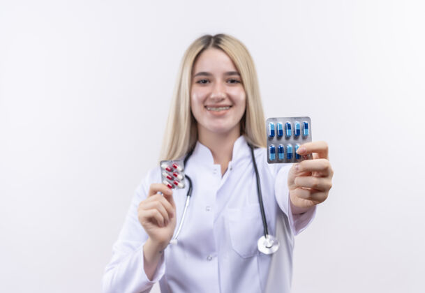 医生微笑的医生年轻的金发女孩戴着听诊器和医用长袍 戴着牙套 双手拿着药片 背景是孤立的白色年轻医疗两者