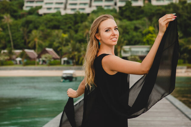 女人穿着黑色连衣裙的美丽迷人的女人在豪华度假酒店的码头上摆姿势 暑假 热带海滩放松挥舞优雅