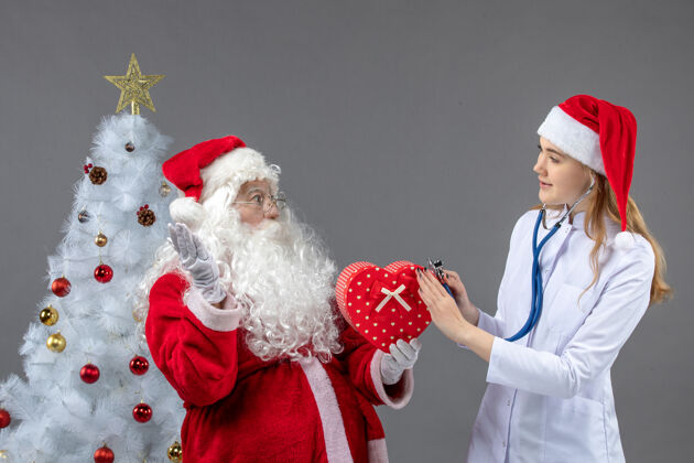 人圣诞老人和女医生的前视图 女医生在灰色墙上用听诊器观察礼物圣诞老人节日礼物
