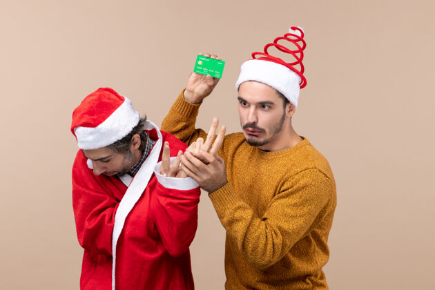 外套前视图两个困惑的家伙一个穿着圣诞老人外套看着地板 另一个拿着米色隔离背景的卡片迷茫拿着两个