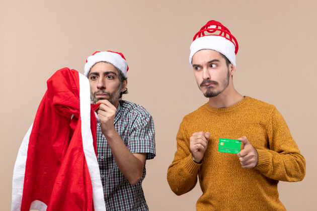 男人前视图两个家伙一个看着圣诞老人的外套 另一个拿着信用卡在米色孤立的背景下看着他伙计们两个前面