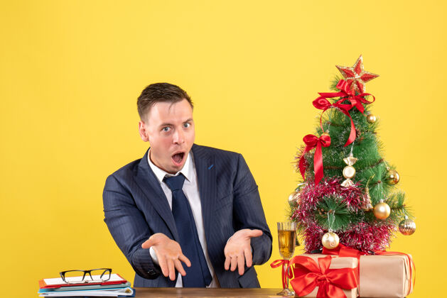 好奇的男人坐在圣诞树旁桌子旁的一个好奇的人 把礼物放在黄色的桌子上会议男性黄色