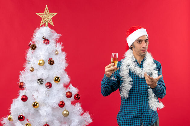 白圣诞节心情与好奇的年轻人圣诞老人帽子在一个蓝色的衬衫剥离举起一杯葡萄酒附近圣诞树圣诞装饰年