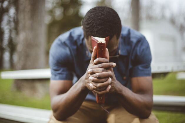 阅读年轻的非洲裔美国男子坐着闭着眼睛 手里拿着圣经一起美国人书