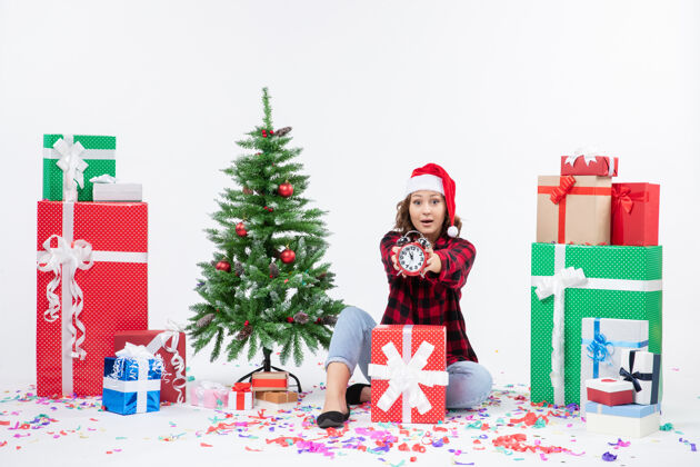 快乐前视图的年轻女子坐在圣诞礼物周围拿着白色墙上的时钟坐着装饰十二月