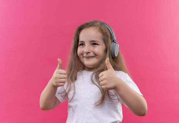 微笑微笑的小女孩穿着白色t恤 戴着耳机听音乐 在孤立的粉色背景下竖起大拇指白色女孩戴着