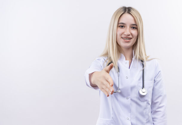 听诊器微笑的医生 年轻的金发女孩 戴着听诊器 穿着医用长袍 戴着牙套 在孤立的白色背景下 向镜头伸出手来医疗白色医生