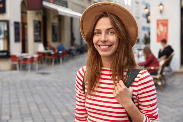 背包可爱的女摄影师喜笑颜开 戴着帽子和条纹套头衫 心情舒畅地漫步在城市中人类城市高兴