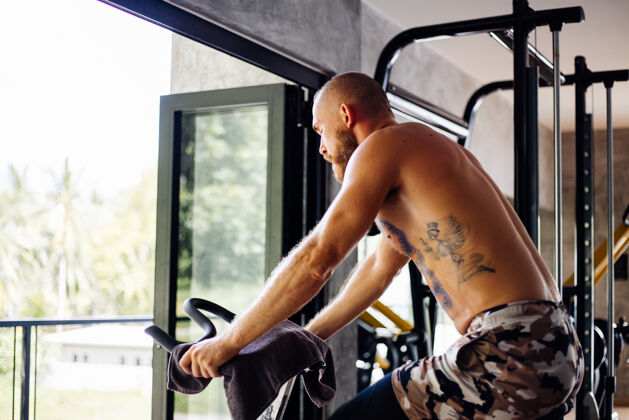 锻炼纹身 肌肉发达 胡须浓密的男人骑着自行车在体育馆里锻炼有氧运动 靠近大窗户 外面可以看到树训练骑自行车男人