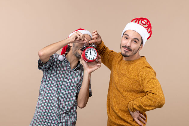 男性前视图两个圣诞节男子拿着红色闹钟在米色孤立的背景米色男人闹钟