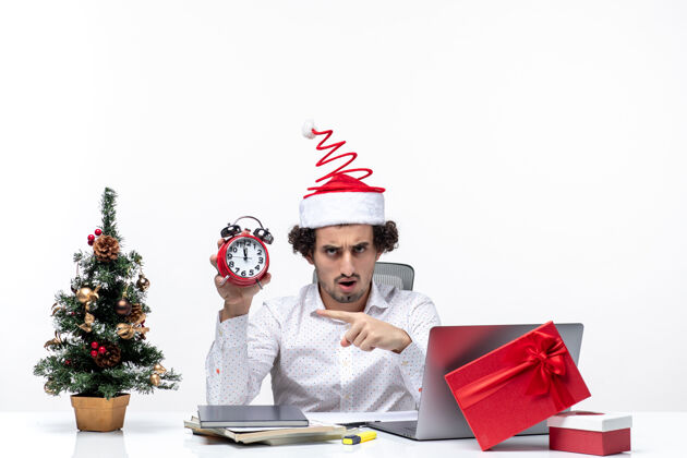 人戴着圣诞老人帽 拿着钟 在白色背景上指着办公室里的东西的年轻商人商务时钟孩子
