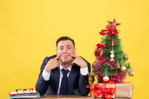 行政人员年轻人坐在圣诞树旁的桌子旁 试图微笑 并在黄色的礼物办公室树坐着的