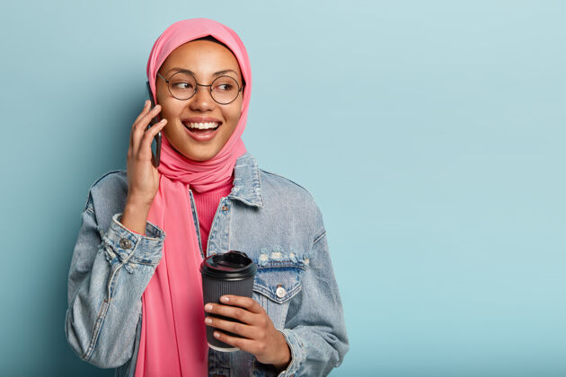 伊斯兰迷人开朗的阿拉伯女人与密友通电话 端着一次性咖啡 目光移开科技手机阿拉伯语