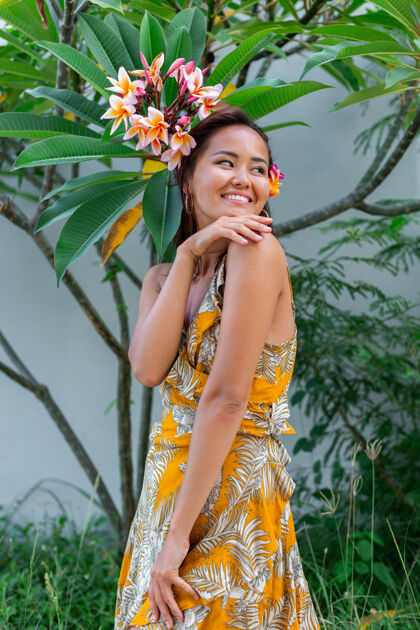 圆形身着黄色夏装的亚洲女性肖像站在墙上和绿色灌木丛的背景上 头发上有一朵鸡蛋花泰国花 耳朵圆圆的女性在外面化妆热带生活方式户外