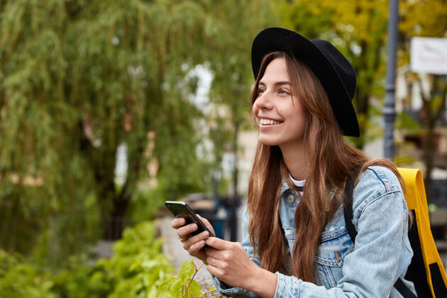 女士开心少女横拍通过手机与朋友聊天 在小玩意上安装应用程序 戴上时尚的黑帽子信息头饰背包
