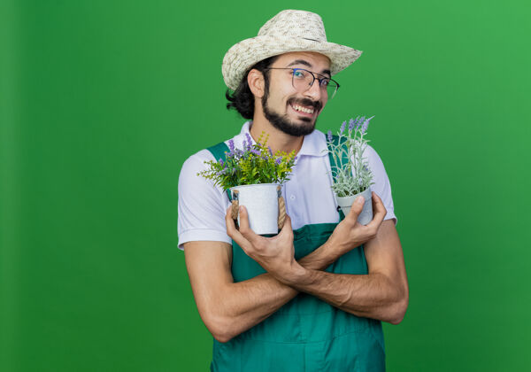 绿色年轻的留着胡子的园丁 穿着连体衣 戴着帽子 手里拿着盆栽植物 满脸笑容看男人胡须