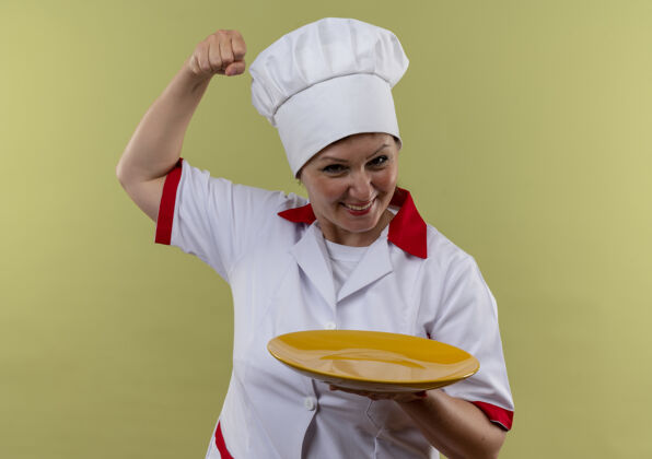 制服笑容可掬的中年女厨师穿着厨师制服拿着盘子做着有力的手势 带着临摹的空间盘子厨师做着