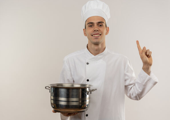 手指面带微笑的年轻男厨师穿着厨师制服拿着平底锅用手指向上抄起空间厨师点男