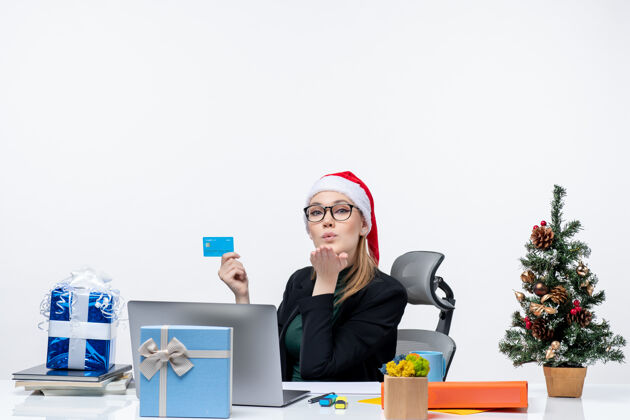 眼镜自信迷人的女人 戴着圣诞老人的帽子 戴着眼镜 坐在桌子旁 手里拿着银行卡 在办公室里送吻亲吻圣诞老人发送
