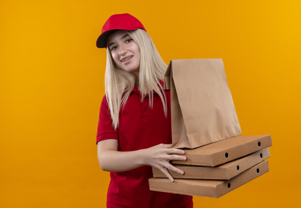 牙科微笑送货的年轻女孩穿着红色t恤 戴着帽子 戴着牙套 拿着披萨盒和纸口袋 背景是孤立的橙色女孩拿着戴着