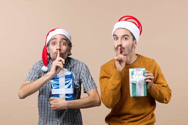 礼物正面图：两个男人在米色的背景上做着嘘的手势 手里拿着圣诞礼物成人肖像伙计们