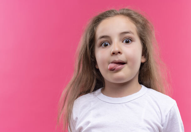 小一个穿着白色t恤的小女孩 在孤立的粉色背景下露出舌头学校穿女孩