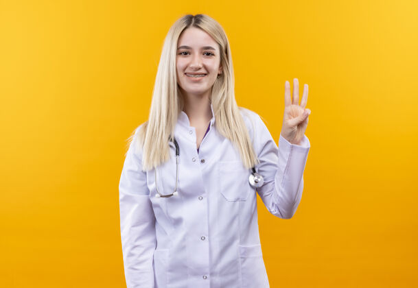 支架微笑的医生年轻女孩穿着医用长袍戴着听诊器和牙套 在孤立的黄色背景上显示三个女孩穿着展示