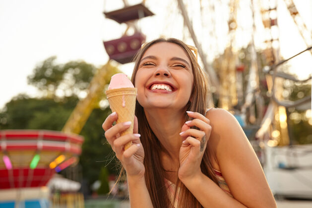 衣服特写镜头：迷人 开朗 棕发的年轻女士头戴墨镜 露出牙齿 开心地微笑着 一边在游乐园吃着粉红色的冰淇淋蛋筒快乐微笑漂亮
