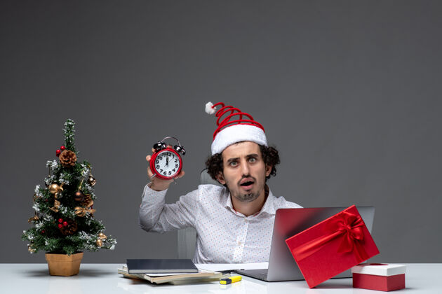 办公室年轻有趣 情绪激动的商务人士戴着圣诞老人的帽子 拿着并展示着黑色背景下坐在办公室里的时钟圣诞老人情绪化微笑