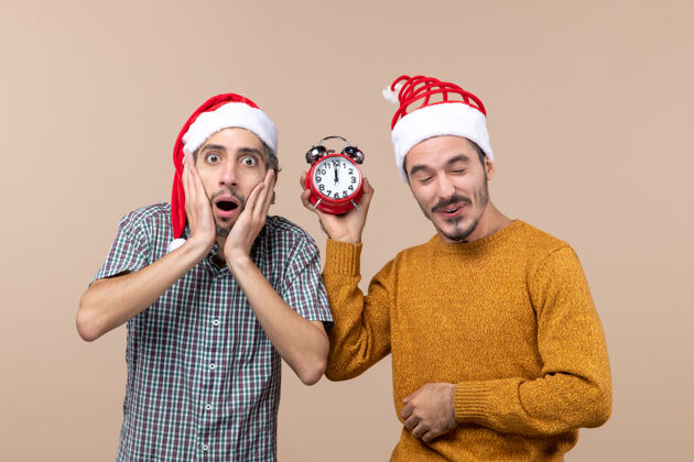 圣诞节前视图两个圣诞节男人一个震惊 另一个拿着闹钟在米色孤立的背景两个圣诞男人前面脸