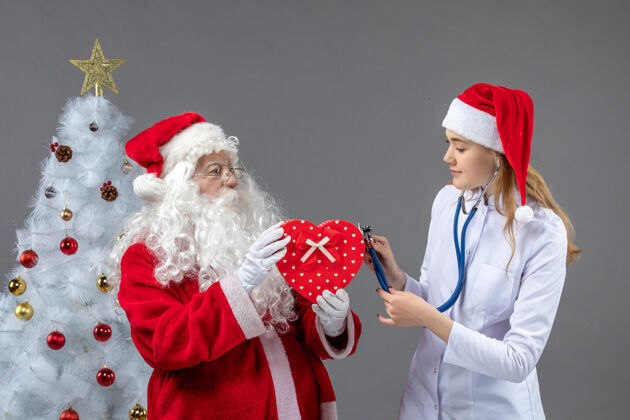 圣诞快乐圣诞老人和女医生在灰色墙上用听诊器观察的正视图男人圣诞老人圣诞