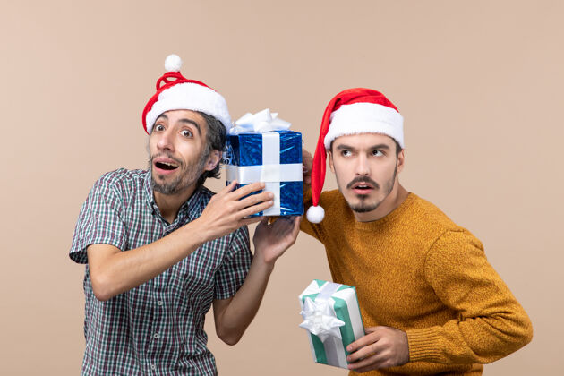 帽子前视图两个戴着圣诞帽的感兴趣的家伙 并注意在米色孤立背景上的圣诞礼物注意圣诞老人男人