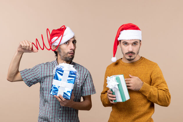 拿着正面图两个戴着圣诞帽的困惑的家伙一个拿着圣诞帽 另一个把手放在米色孤立背景的胸前手男性米色