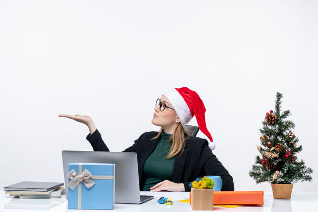 一边一个戴着圣诞老人帽子和眼镜的年轻女子坐在一张桌子旁 桌子上放着圣诞树和礼物 在白色背景上指着右边办公室桌子礼物
