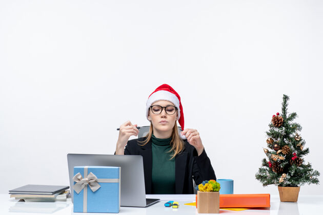 桌子新年的气氛与果断的金发女人与圣诞老人的帽子坐在一张桌子上 圣诞树和一个礼物在它的白色背景办公室圣诞节电脑