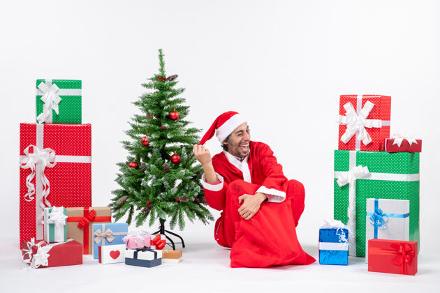 微笑笑容可掬的年轻人打扮成圣诞老人 拿着礼物和装饰好的圣诞树坐在白色背景的地上圣诞老人圣诞圣诞老人