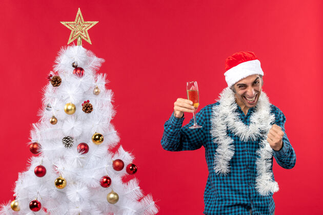 人圣诞心情与骄傲情绪化的年轻人与圣诞树附近拿着一杯葡萄酒的蓝色条纹衬衫圣诞老人帽子年轻庆祝圣诞老人