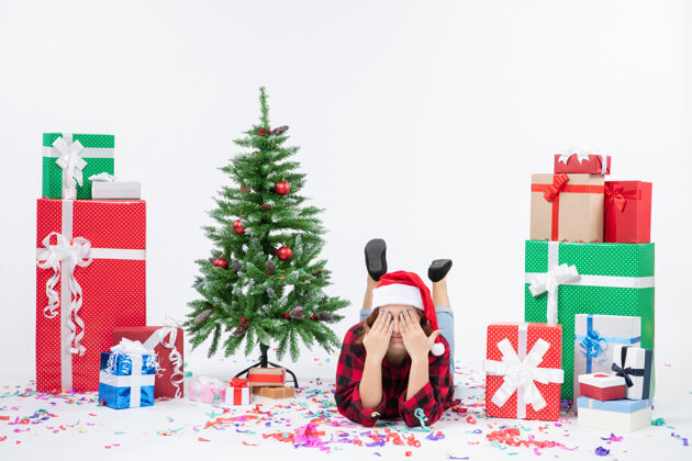 礼物前视图年轻的女性躺在圣诞礼物和小圣诞树周围的白色背景圣诞新年彩雪礼物躺着女性