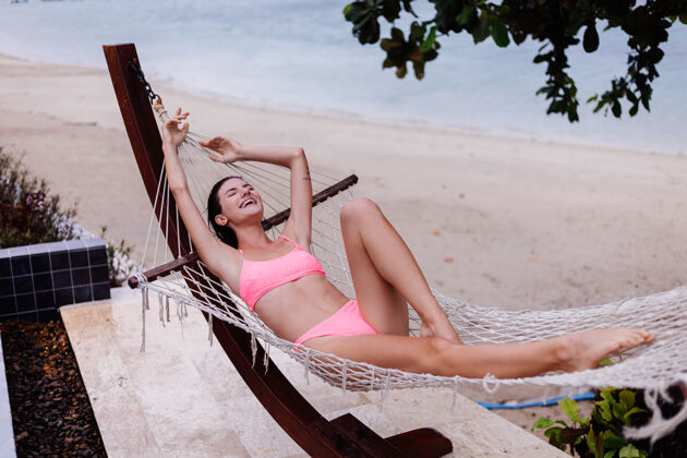 异国情调快乐的白种女人躺在海滩边的吊床上 穿着粉色比基尼健康温暖泰国