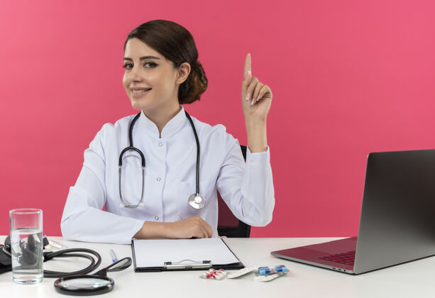 工作面带微笑的年轻女医生穿着医用长袍 手持听诊器坐在办公桌旁 用电脑和医疗工具指着上面的复印空间办公桌医生穿