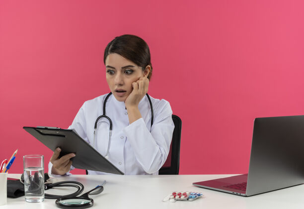 惊讶惊讶的年轻女医生穿着医用长袍 手持听诊器 坐在办公桌旁 拿着医疗工具 拿着电脑 看着有复印空间的剪贴板医疗医生长袍